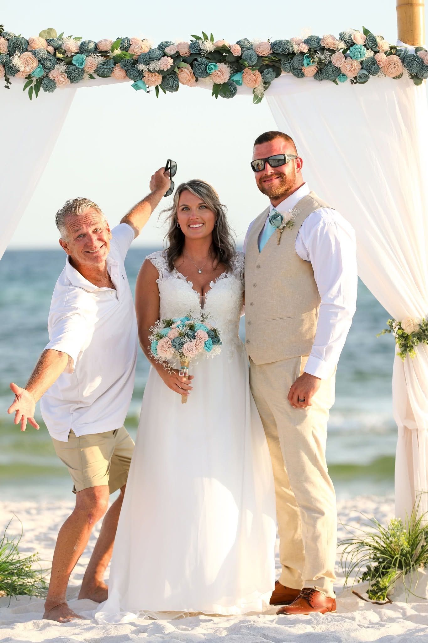 Three people posing under a floral arch on a beach, with one holding a bouquet and all smiling.