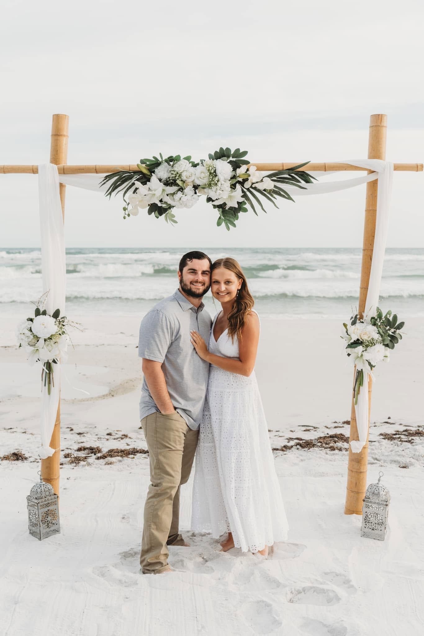 Couple standing on a beach under a floral wedding arch, wearing white and tan, with ocean waves behind them