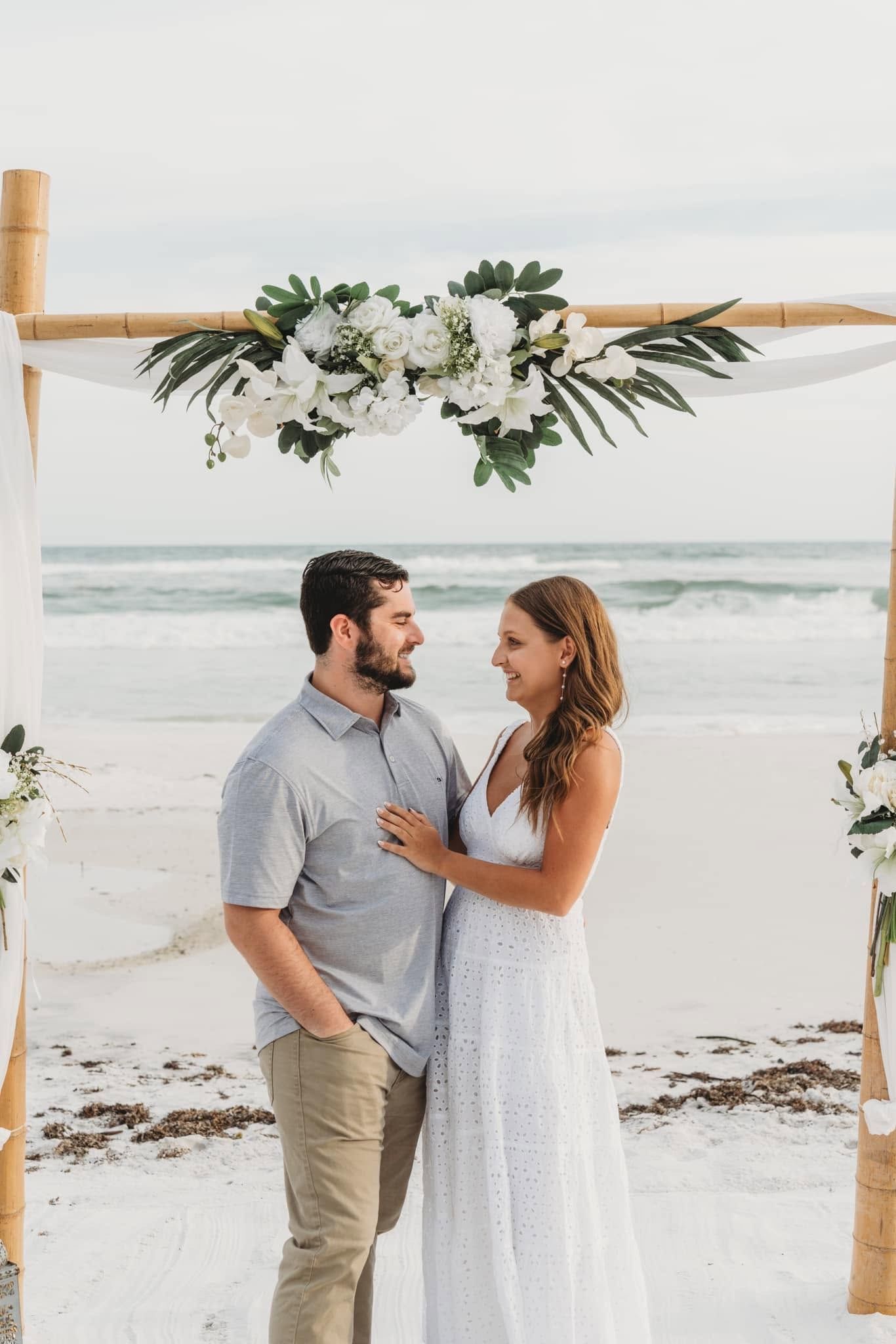 Couple standing under floral arch on a beach at sunset, facing each other and holding hands