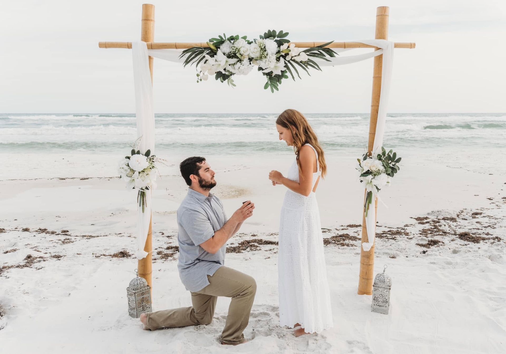 Beach wedding proposal under a flower-adorned arch, with a man kneeling before a woman in a white dress.