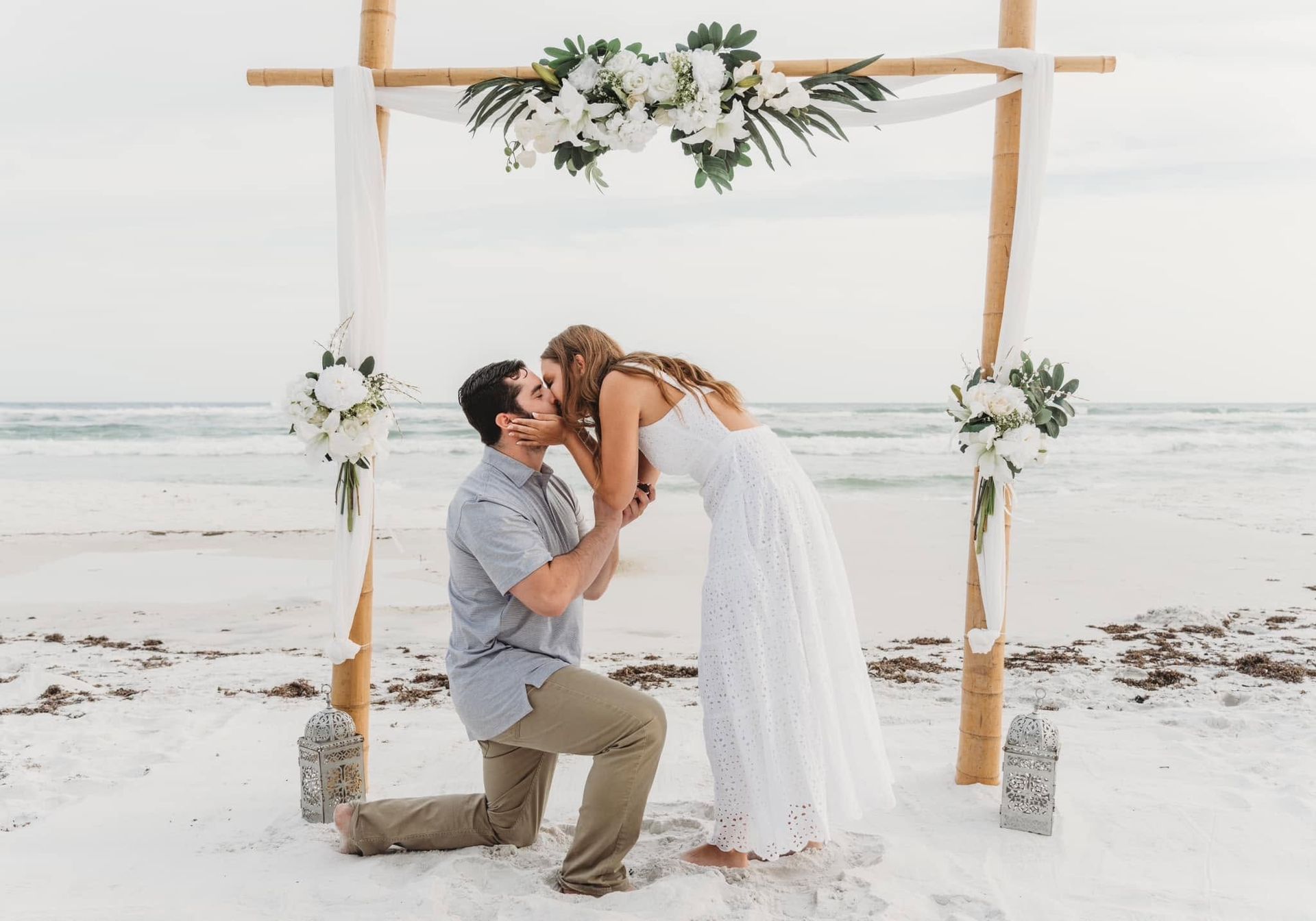 Couple kissing under a floral wedding arch on a white sandy beach at sunset
