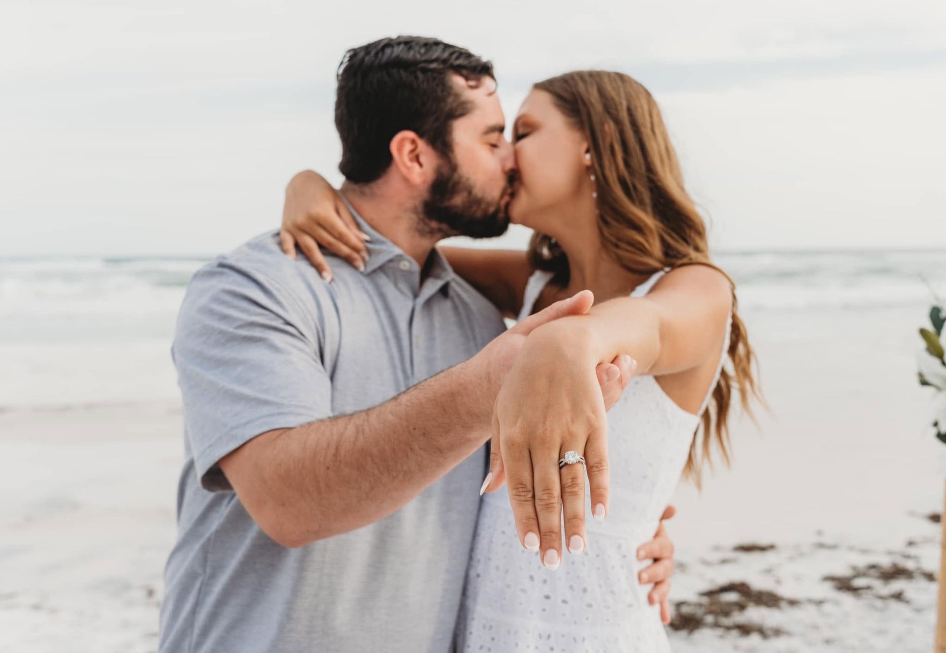 Couple kissing on a beach, with the woman showing an engagement ring.