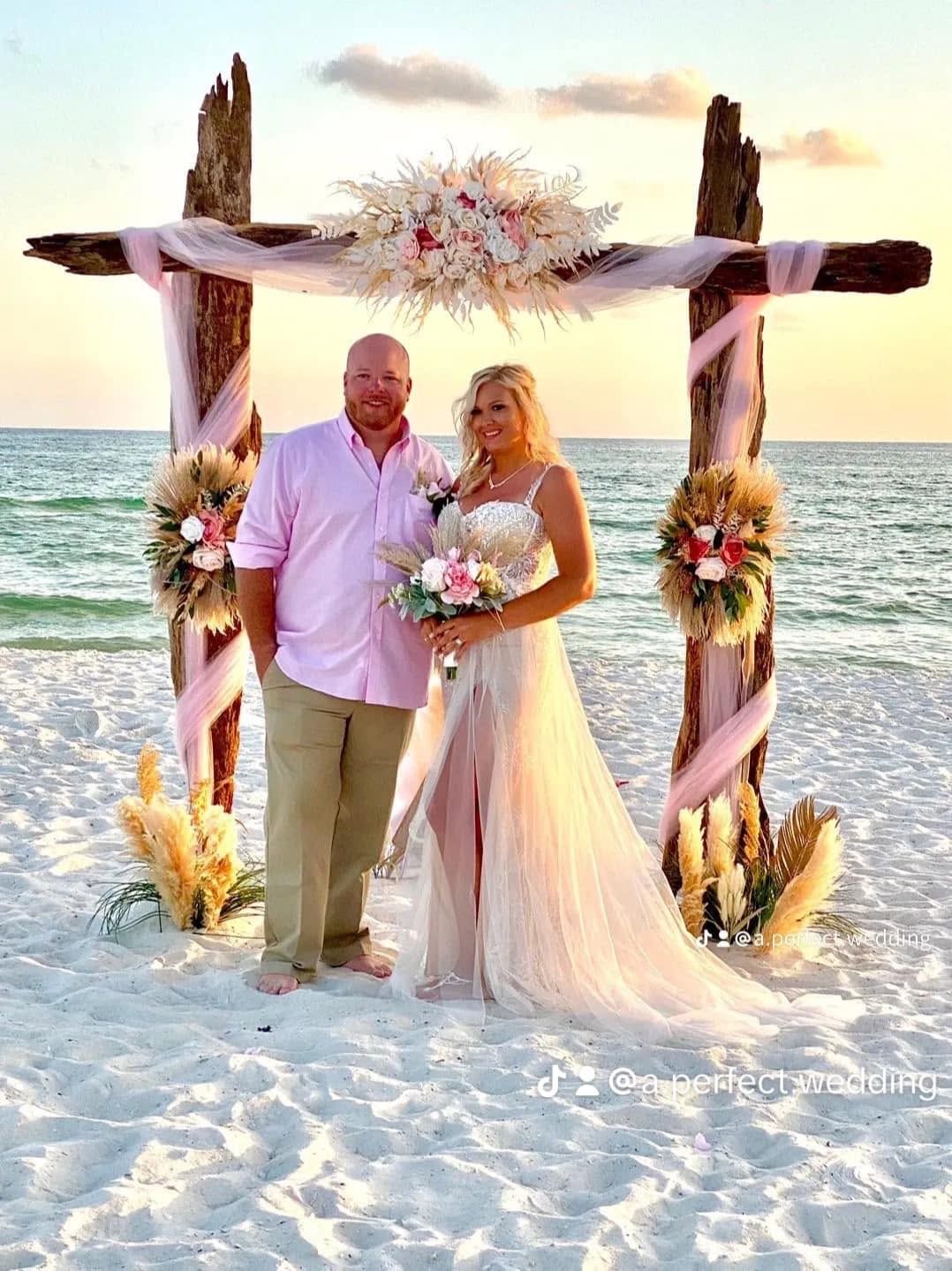 Couple standing under a floral wedding arch on a beach at sunset
