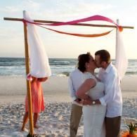 Couple kissing under a white-and-pink beach wedding arch at sunset by the ocean