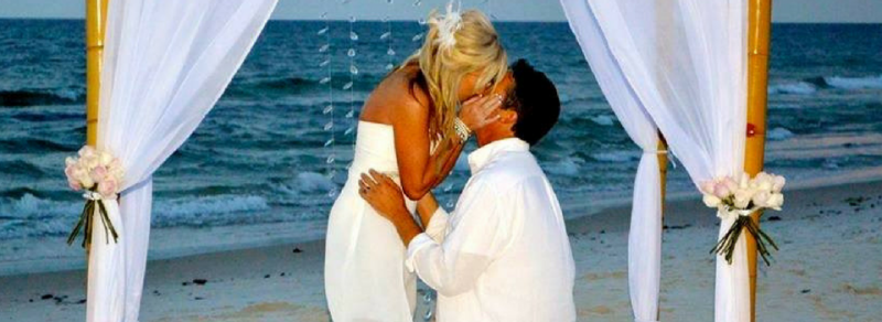 Couple embracing beneath white drapes on a beach wedding arch by the ocean