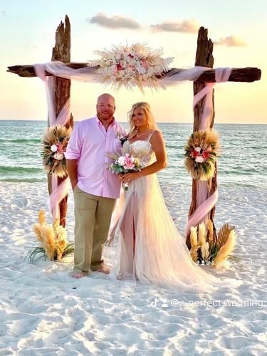 Couple standing under a floral arch on a beach at sunset, with ocean waves behind them.