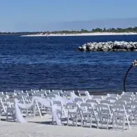 White chairs set on a sandy beach beside blue water and a distant shoreline under a clear sky