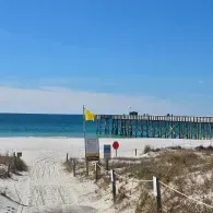 Sandy beach path with dune grass leading to a pier under a bright blue sky and ocean; yellow flag by the walkway
