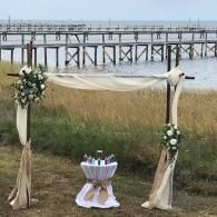 Decorated outdoor wedding arch by the water with draped fabric and white floral arrangements
