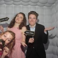 Three people posing with black “love him” signs against a gray backdrop, smiling at a photo booth.
