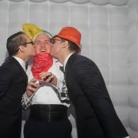 Two men in suits kiss a central person wearing an orange hat and red scarf at a party.