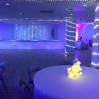 Blue-lit banquet hall with round tables, white tablecloths, and string lights on columns