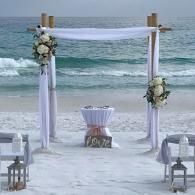 Beach wedding arch with white drapes and floral arrangements by the ocean