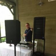 Man standing behind a table with audio equipment beside tall black speakers on a porch