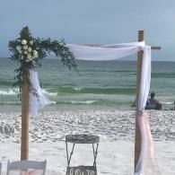 Beach wedding arch with white drapes and floral arrangement by the ocean