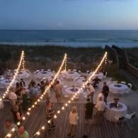 Beachfront dinner party with string lights, round tables, and ocean at dusk
