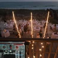 Seaside wedding reception at dusk with round tables, sparkler fountains, and DJ setup in the foreground