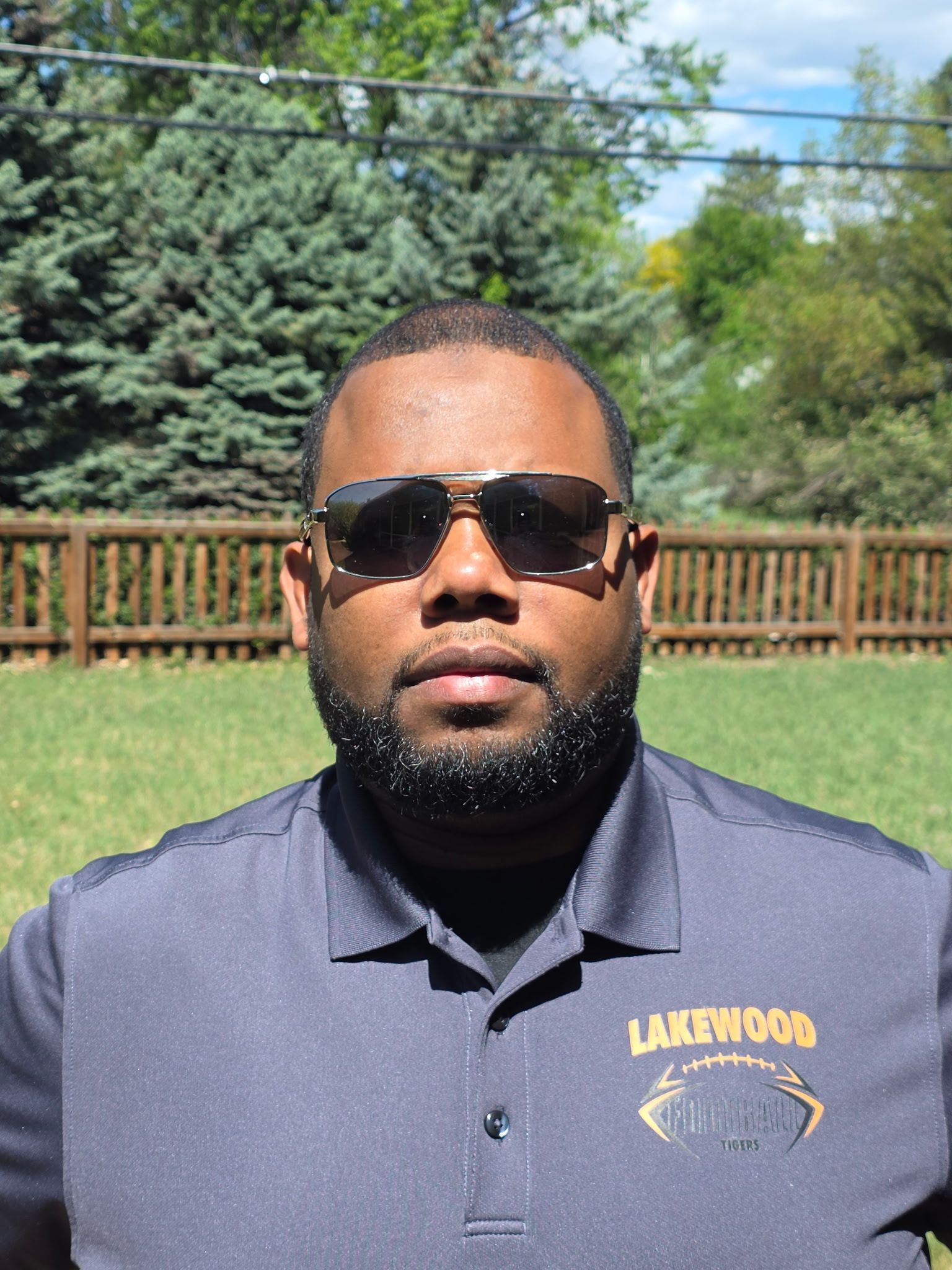 Man wearing sunglasses and a navy polo shirt standing outdoors in front of a wooden fence and trees