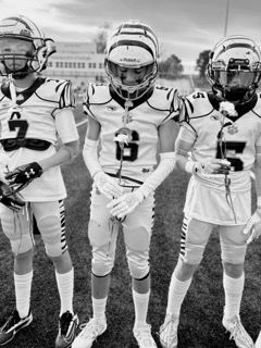 Three young football players in white uniforms and helmets standing on a field, facing the camera.