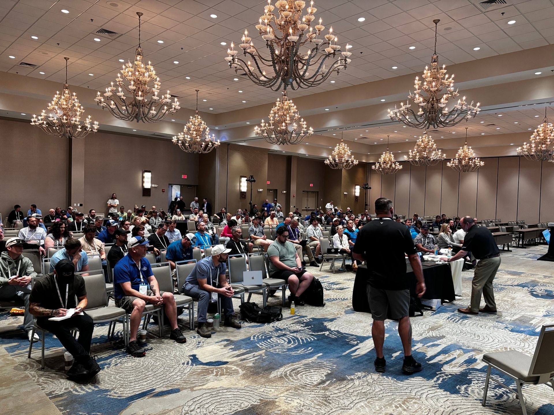 Conference audience seated in a ballroom, facing a speaker under chandeliers.