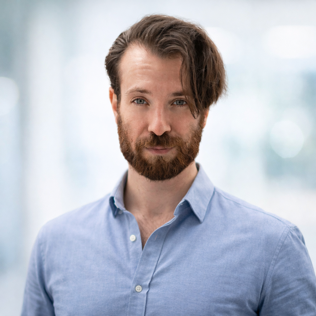 Man with dark hair and beard, wearing a light-colored button-down shirt, looking directly at the camera.
