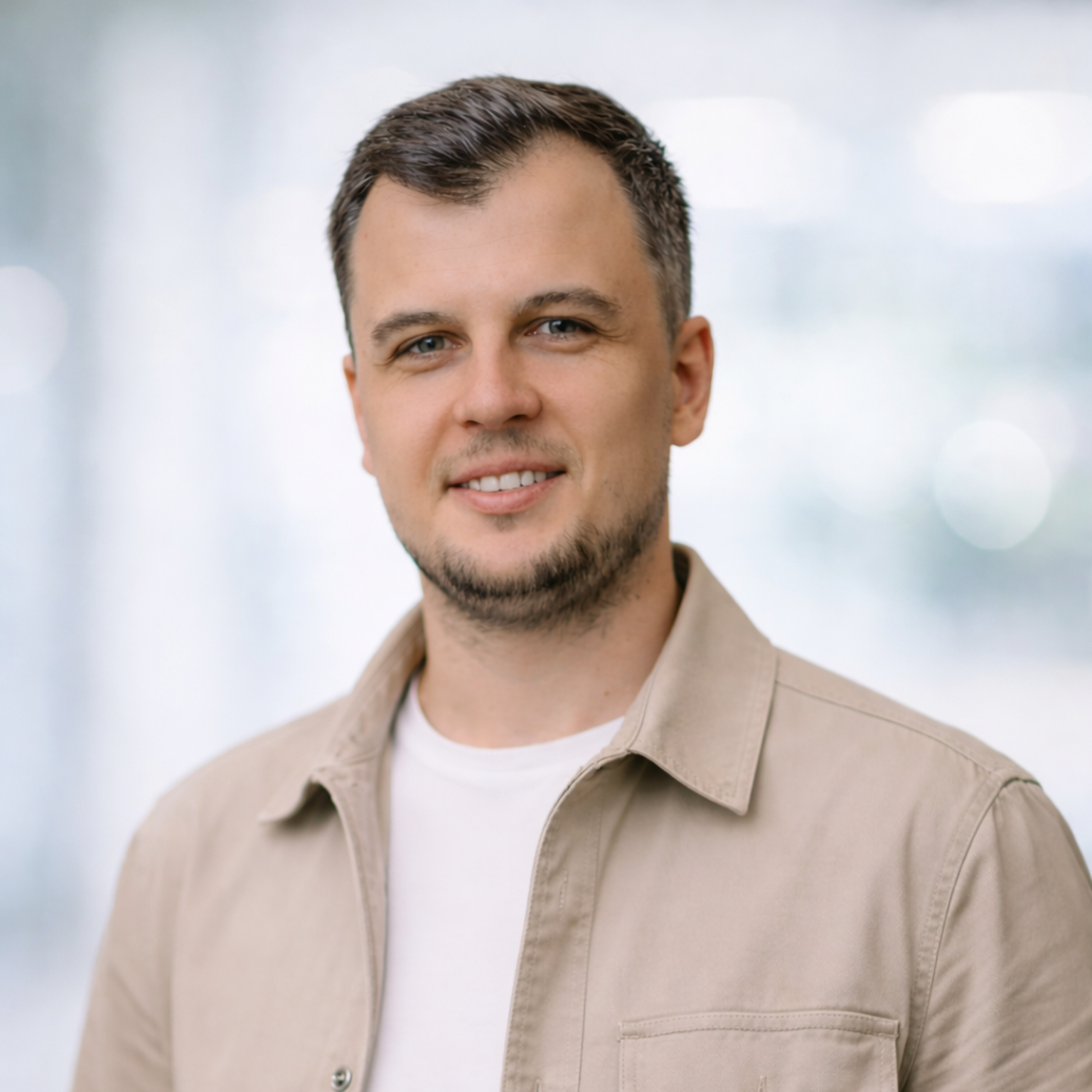 Man with short hair and a small beard smiling, wearing a light-colored button-up shirt over a white tee.