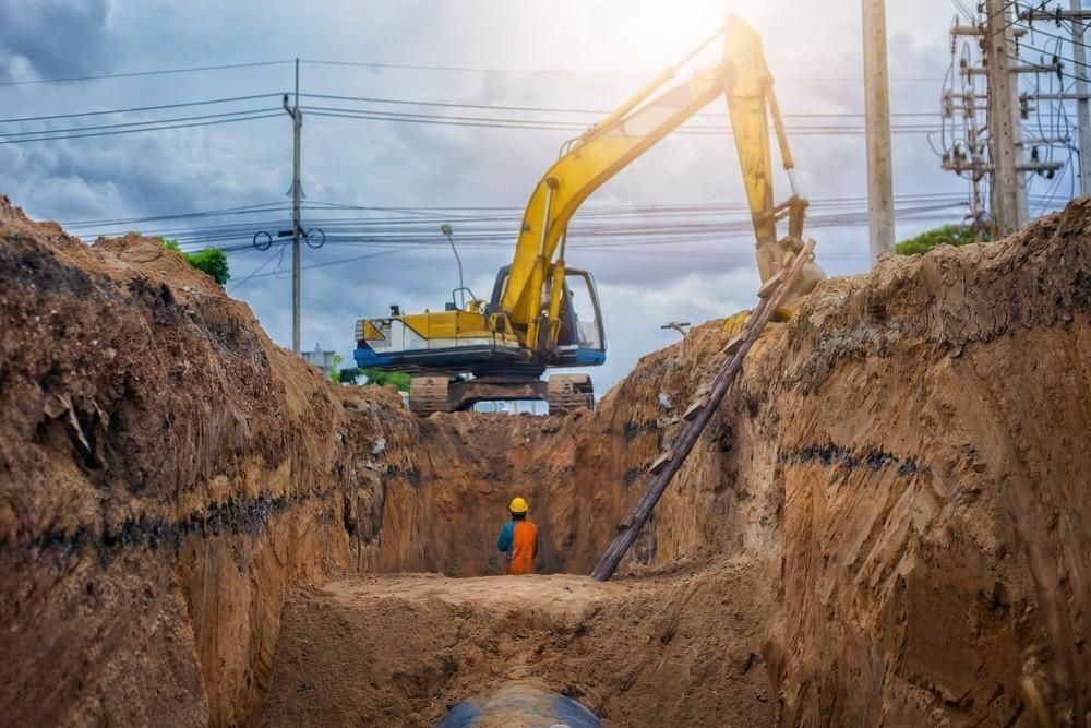 A construction worker is standing in a trench next to a yellow excavator.