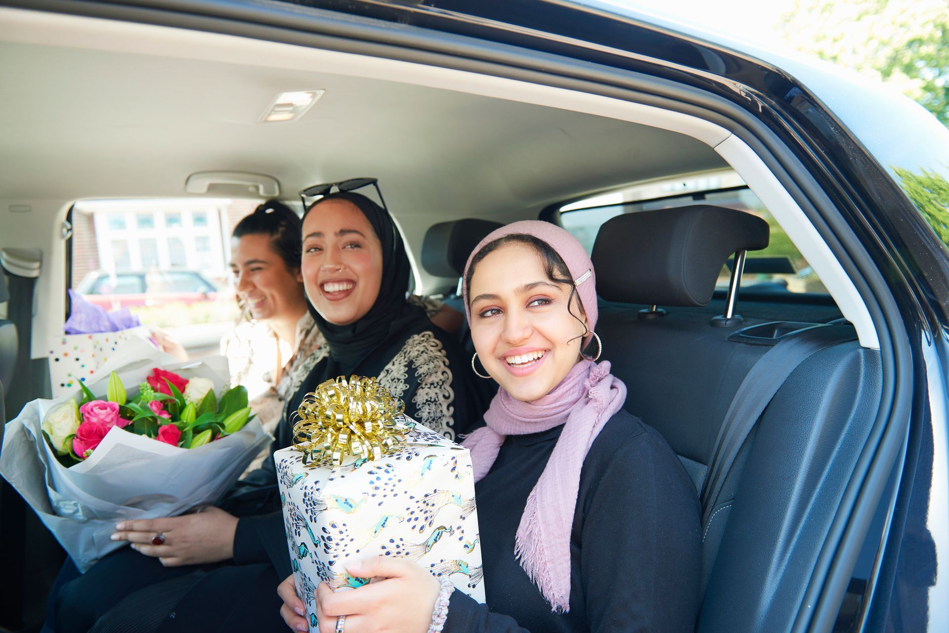 Three women are sitting in the back seat of a car holding flowers and presents. Three women are sitting in the back seat of a car holding flowers and presents.