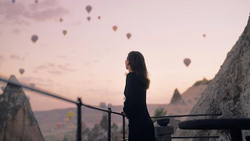 A woman is standing on a balcony looking at hot air balloons flying in the sky.