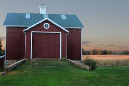 Red barn with closed doors on a grassy hill, overlooking a field at dusk.