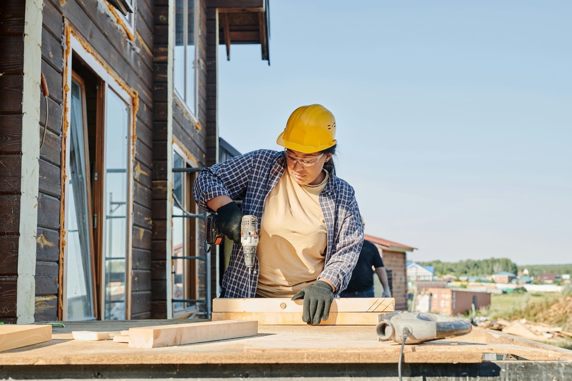 Woman in yellow hard hat drills wood outdoors; building construction.