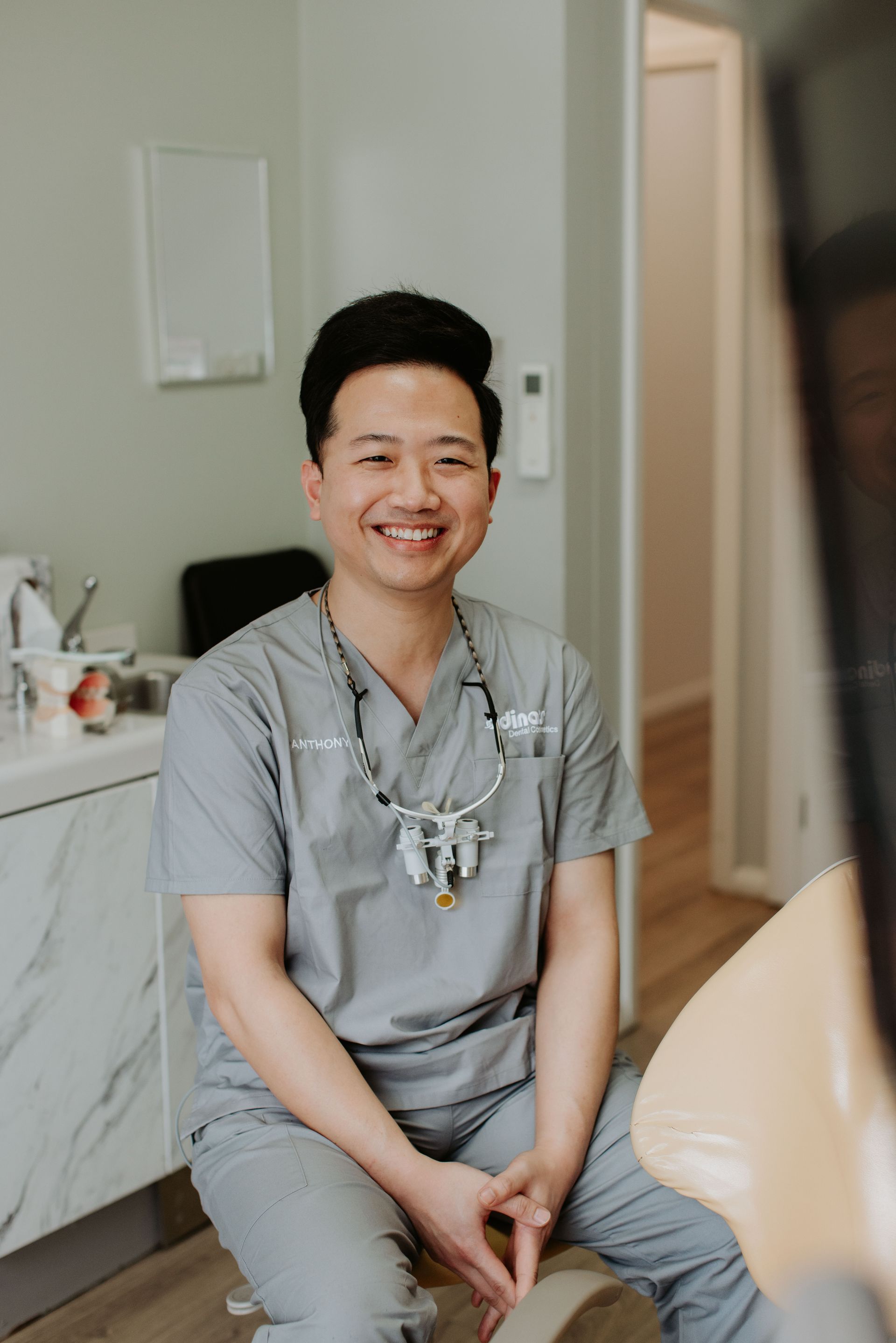A dentist is sitting on a chair in a dental office and smiling.