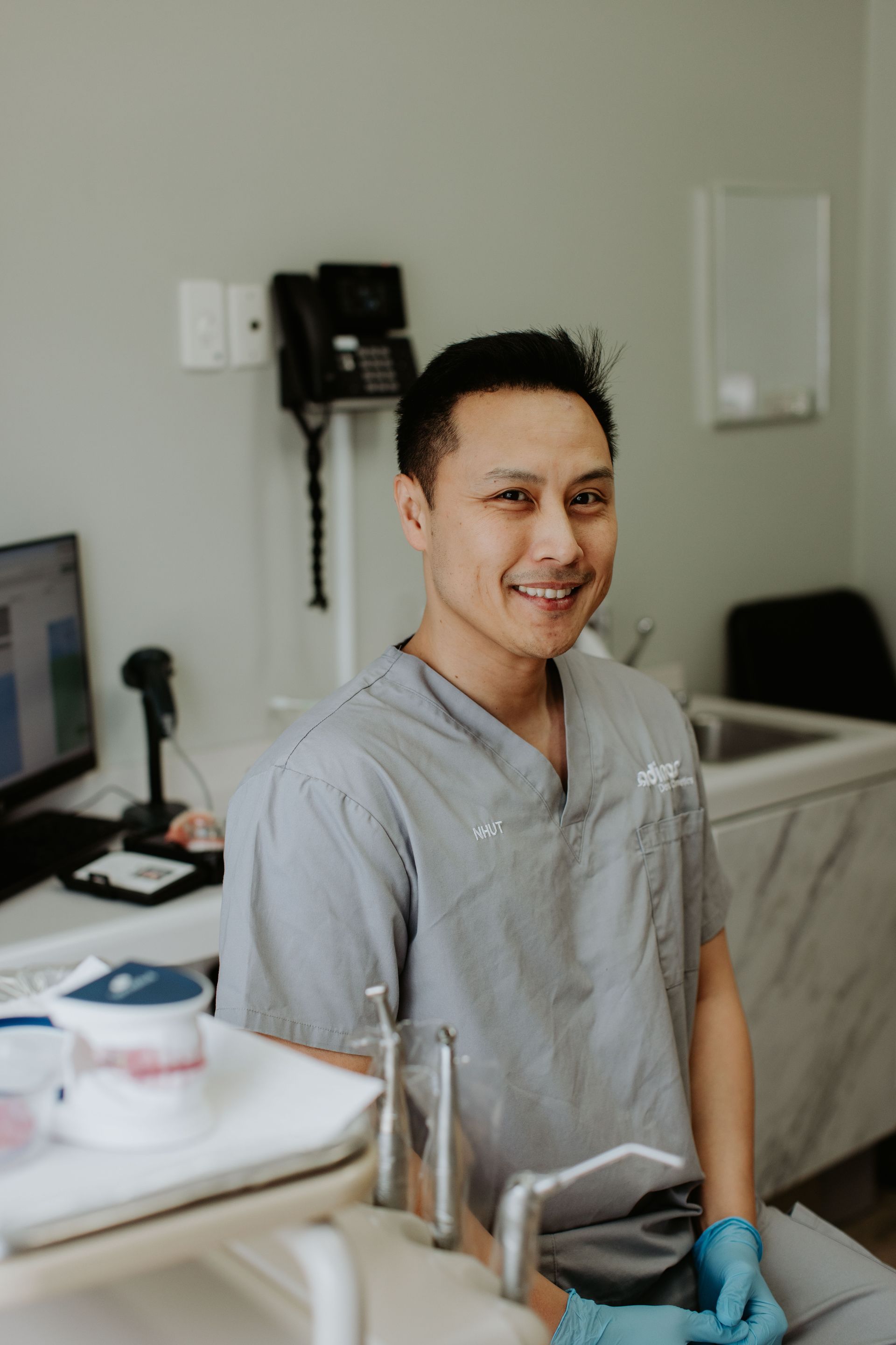 A dentist is smiling while sitting in a chair in a dental office.
