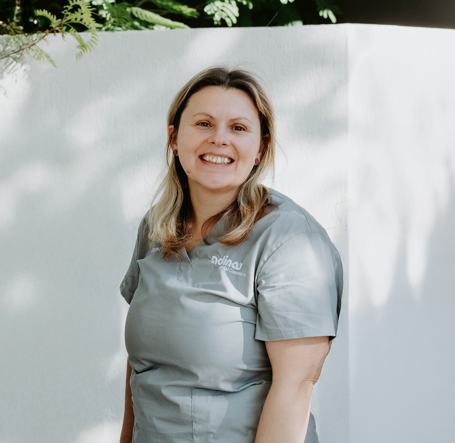 A woman in a grey scrub top is smiling in front of a white wall.