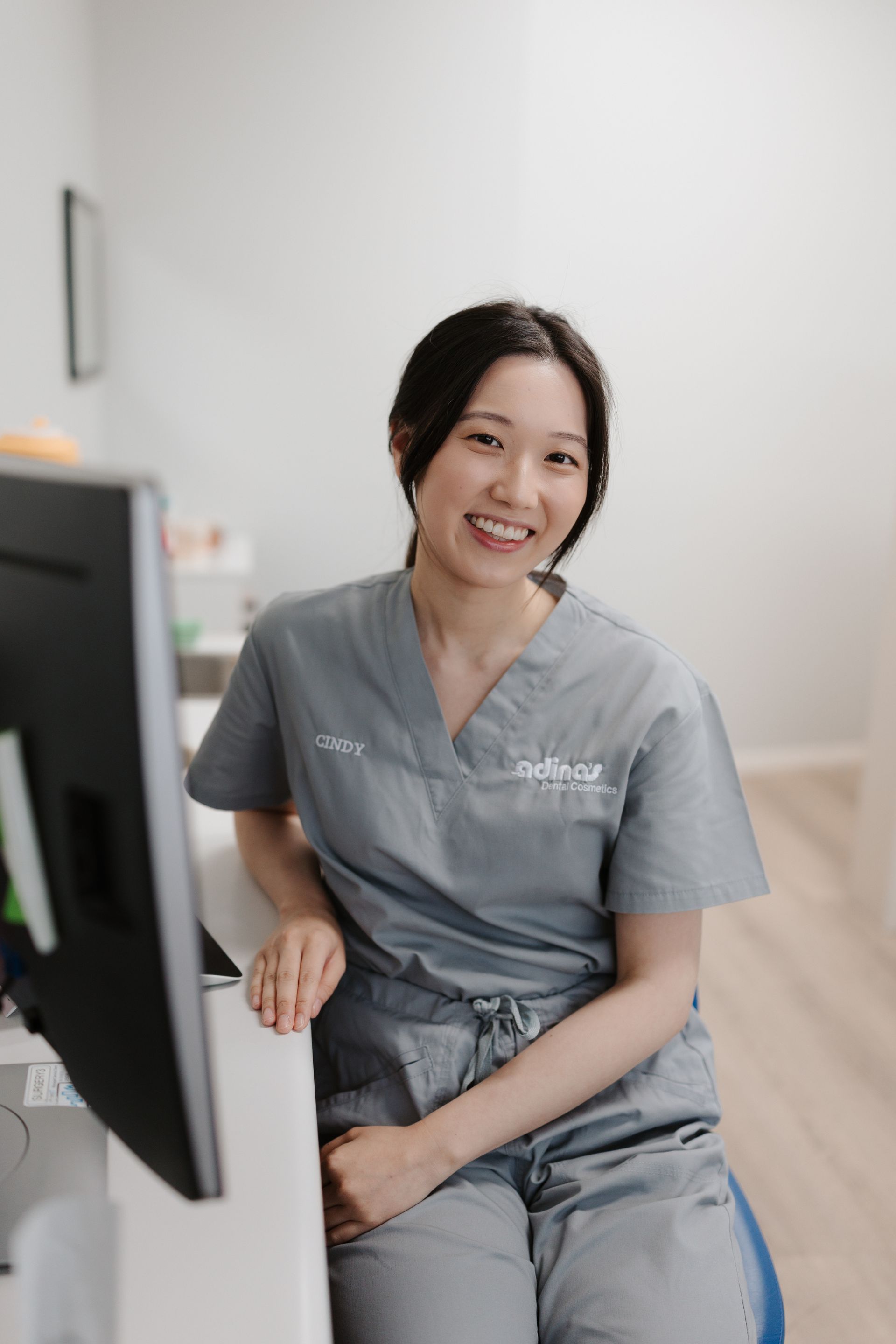 Woman in grey scrubs smiles, leaning on a desk, pin a dental office.