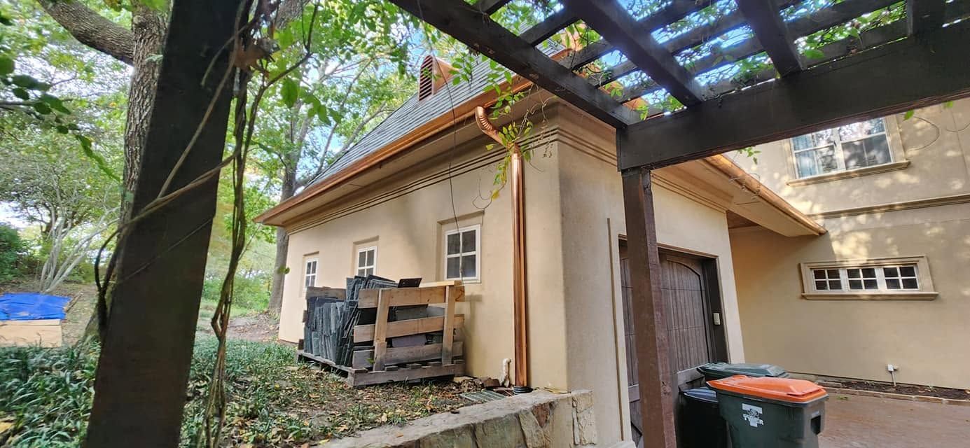 Back patio with a wooden pergola, tan house wall, potted plant, and lush trees nearby.