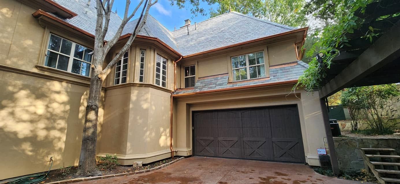 Tan stucco two-story house with dark garage doors, blue roof, and front steps under trees