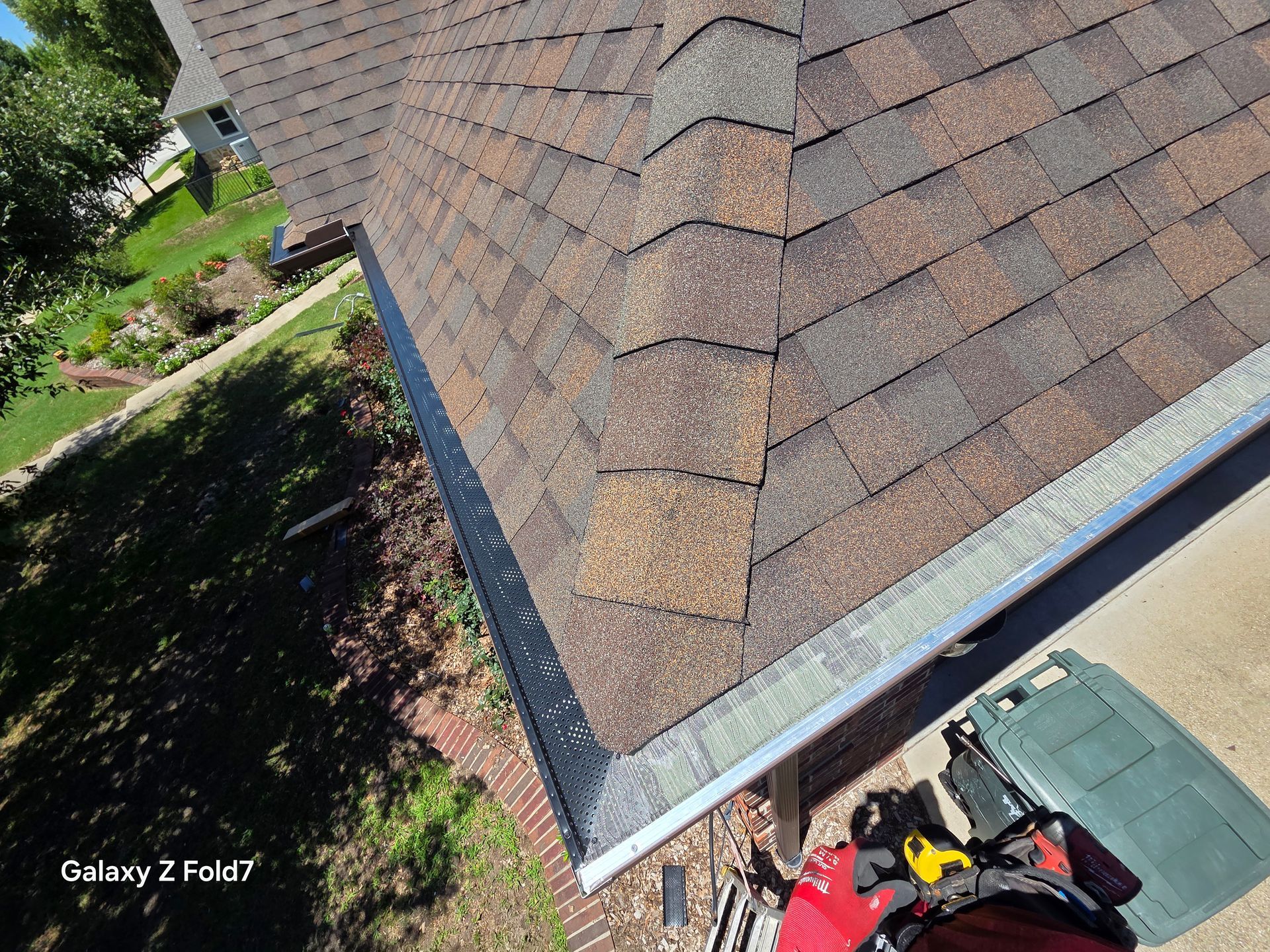 Roof shingles and gutter edge with a person below on a driveway beside a house.