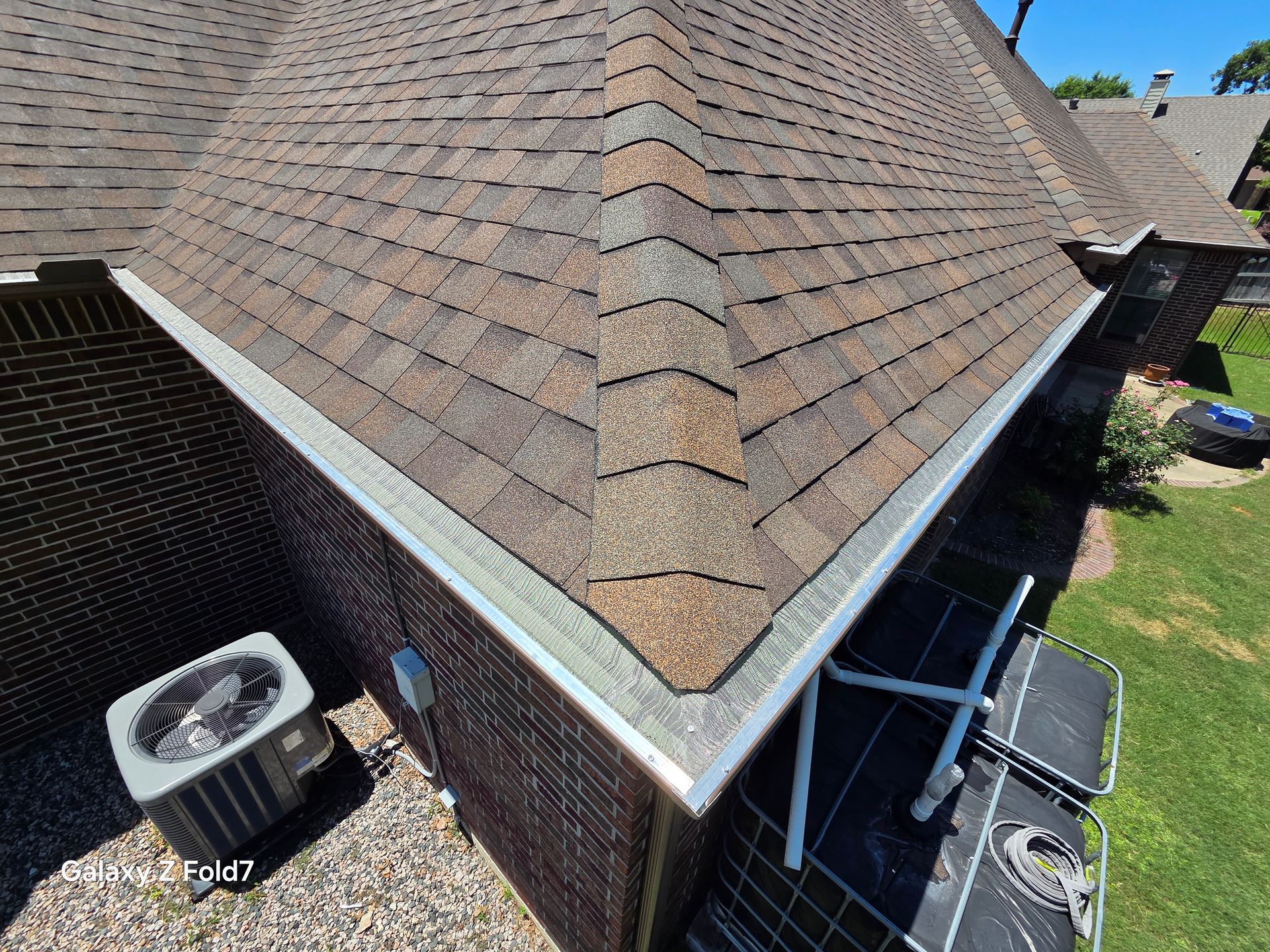 Roof corner with brown shingles and brick siding, showing an HVAC unit and adjacent deck area below.