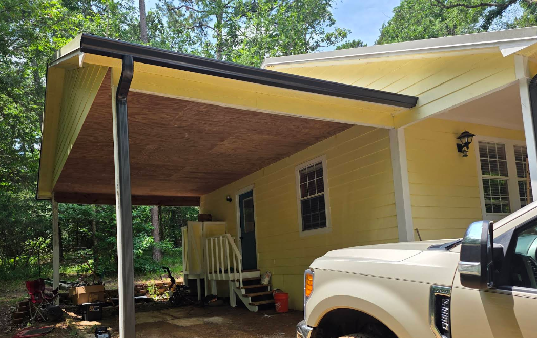 Yellow house with a large covered porch and a white truck parked beside it, surrounded by trees.