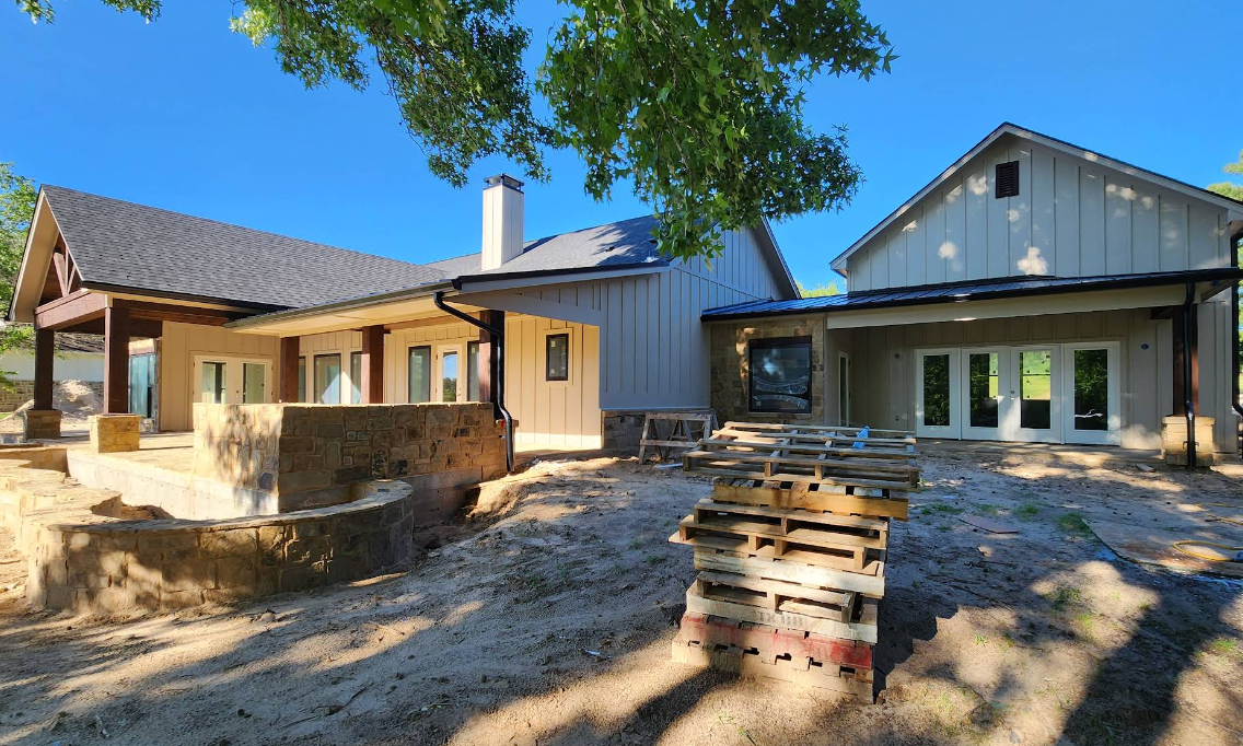 Modern house under construction with stone walls, wooden deck, and stacked lumber in a sunny yard.
