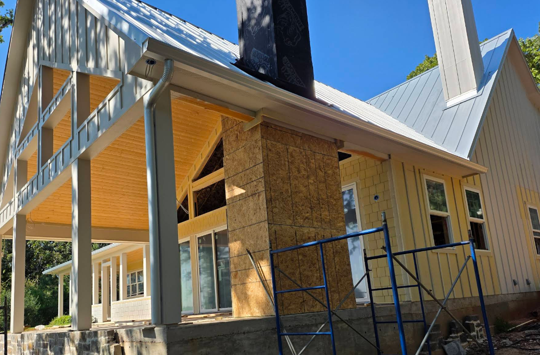 House under construction with exposed wood framing, white siding, and blue scaffolding under a clear sky