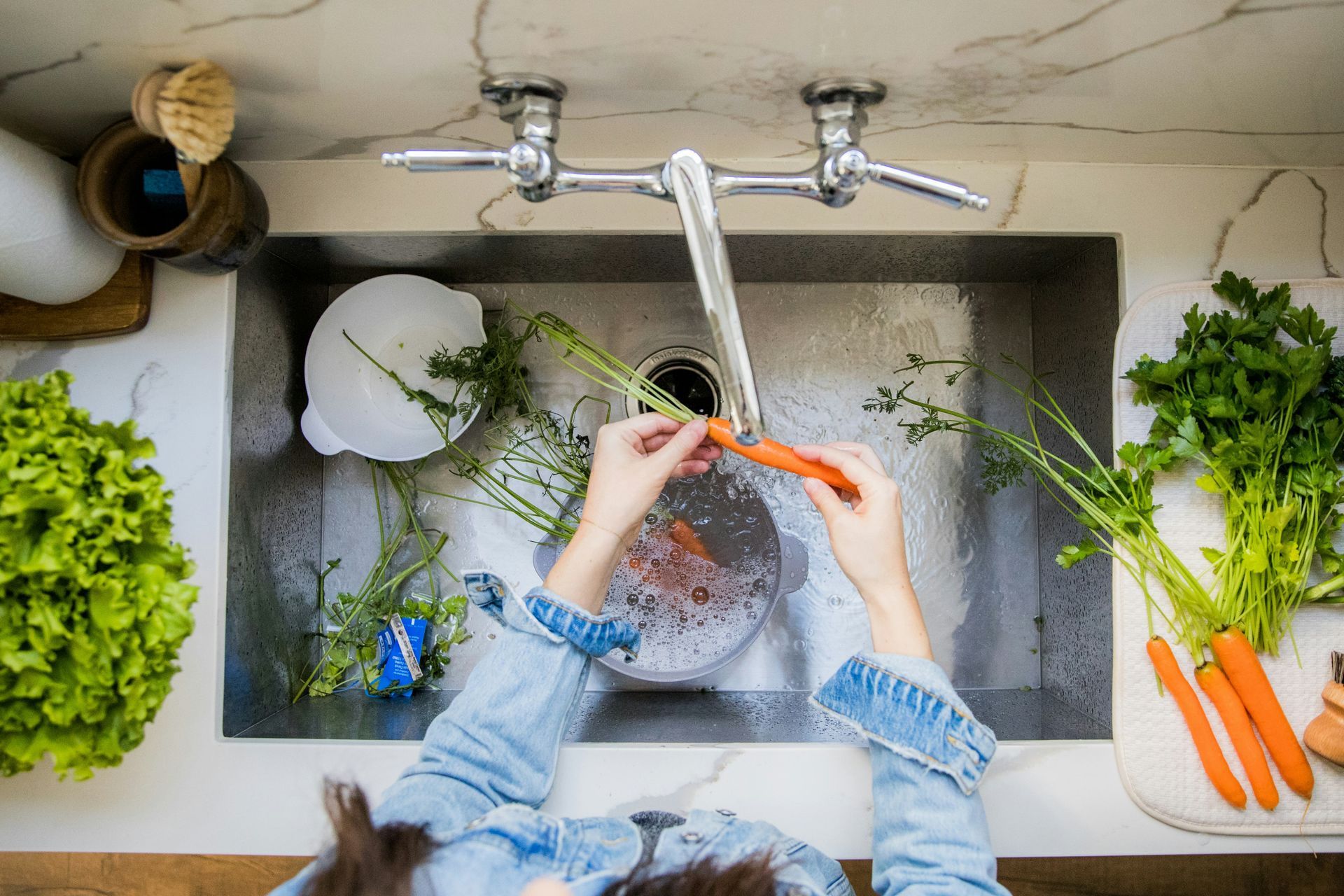 Woman washing dishes with older woman in a kitchen; smiling, wooden shelves, white cabinets.