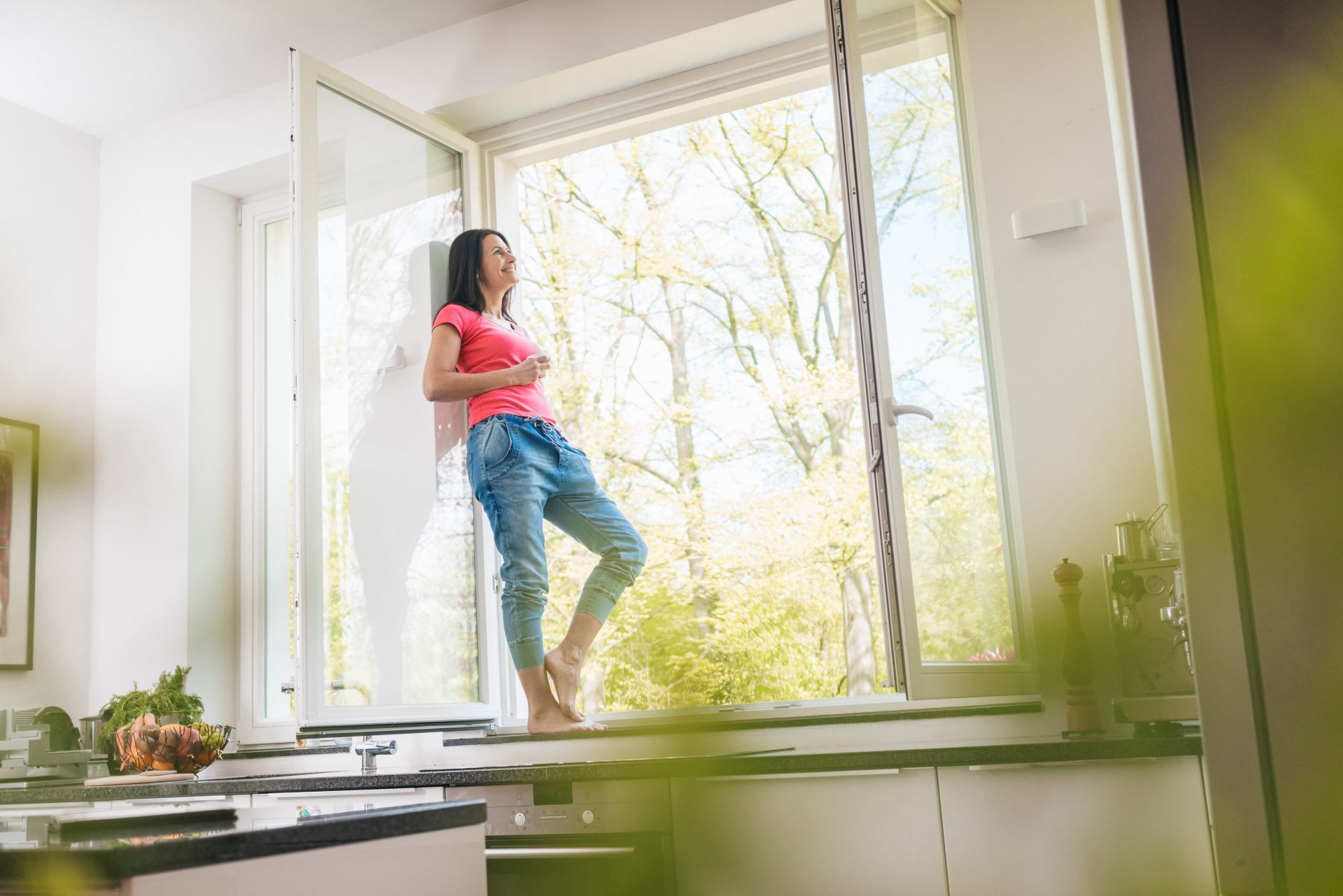 A woman is standing on a window sill in a kitchen looking out the window.