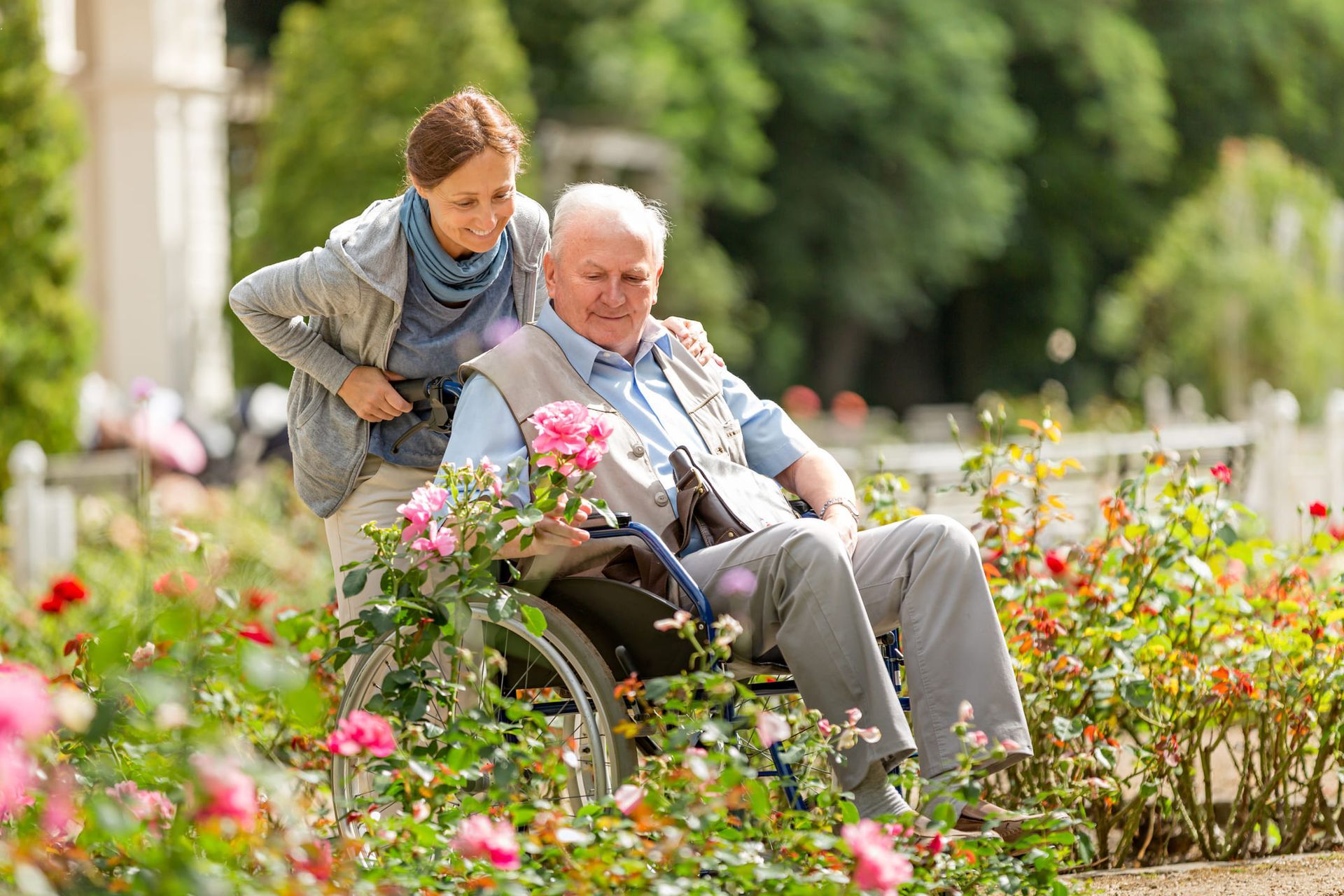 A woman is smiling while pushing an old man in a wheelchair in a garden