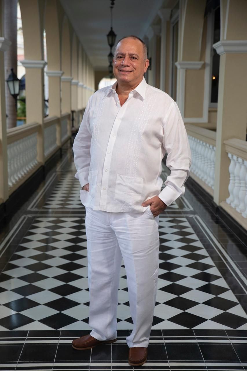 Un hombre con camisa blanca y pantalones blancos está parado en un pasillo con las manos en los bolsillos.