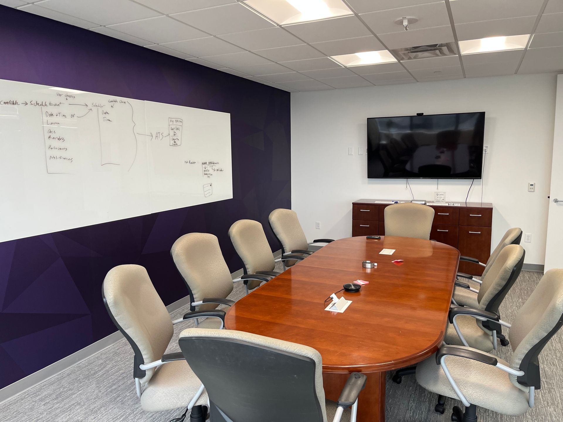 Conference room with oval table, chairs, whiteboard, TV, purple wall.