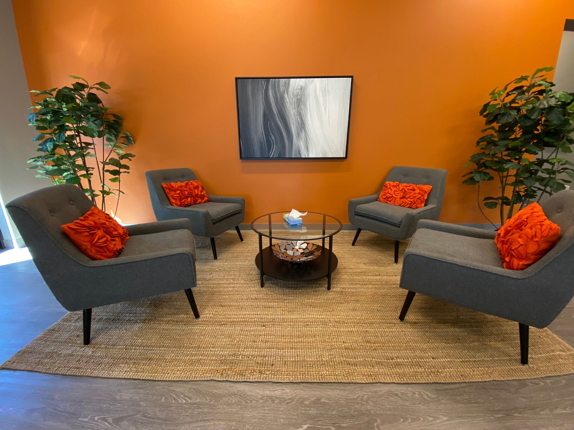 Four gray armchairs with orange pillows around a coffee table on a rug, in front of an orange wall with art.