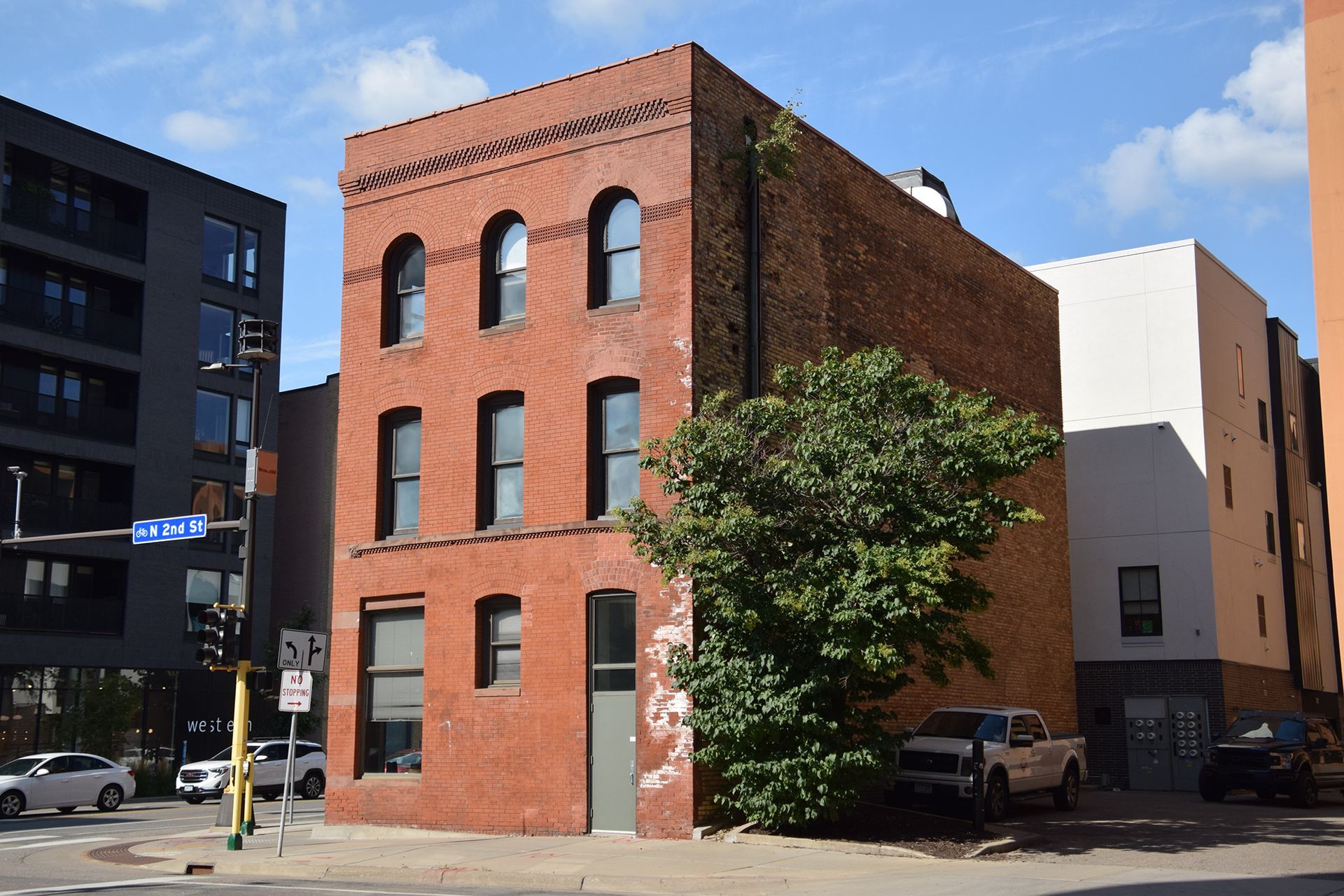 Three-story red brick building on a city street corner, with a modern black building and white building next to it.