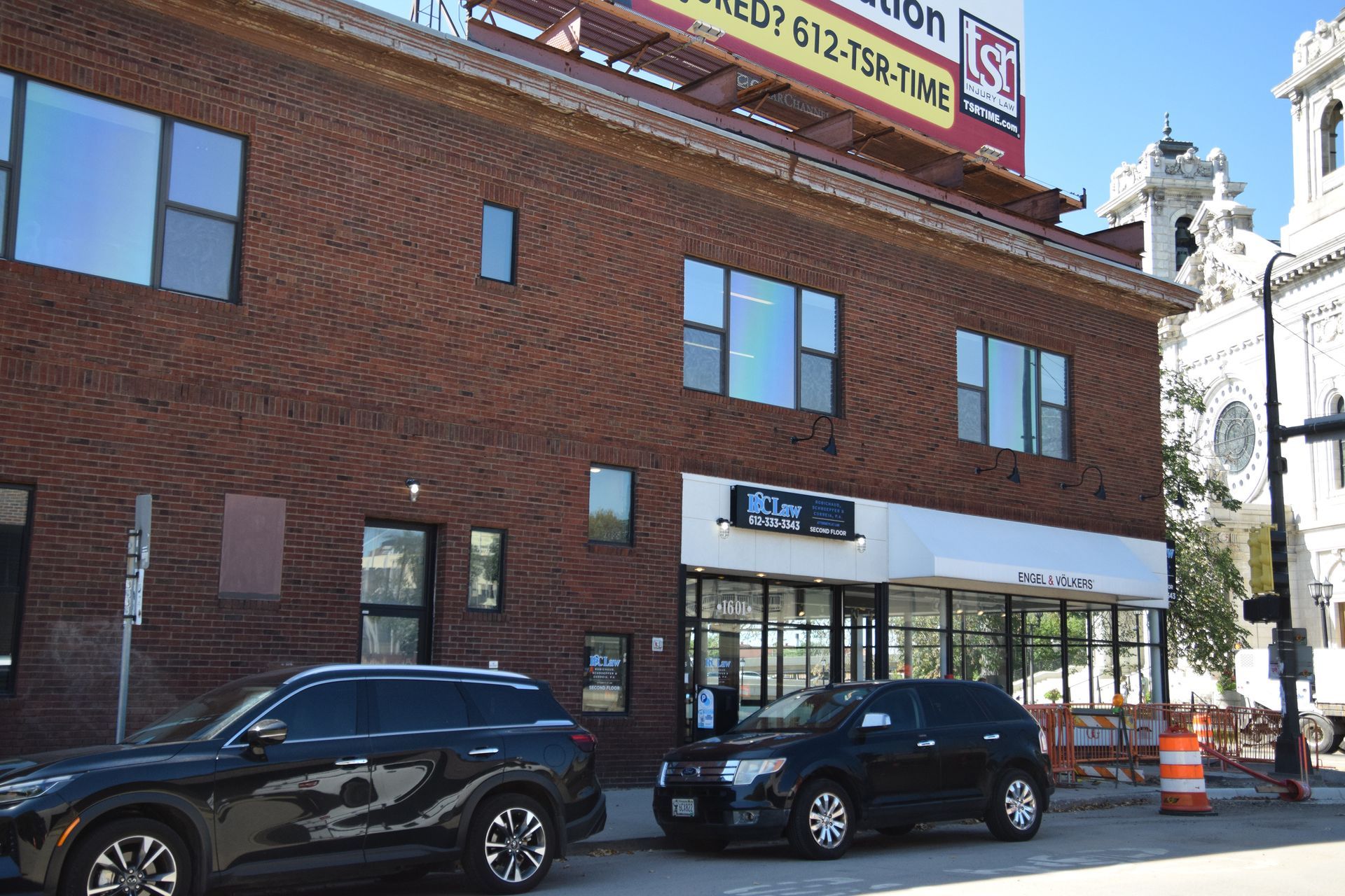 Brick building with several windows, sign over glass door, cars parked in front.